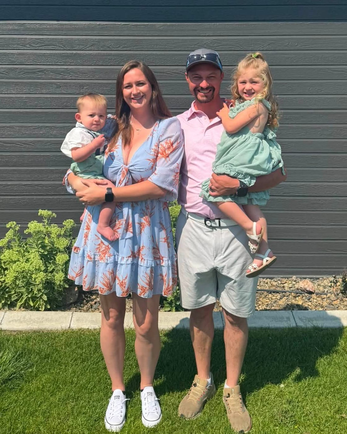 Mitch and Kaitlyn standing in front of a dark-sided building, Kaitlyn holding their baby son and Mitch holding their young daughter.