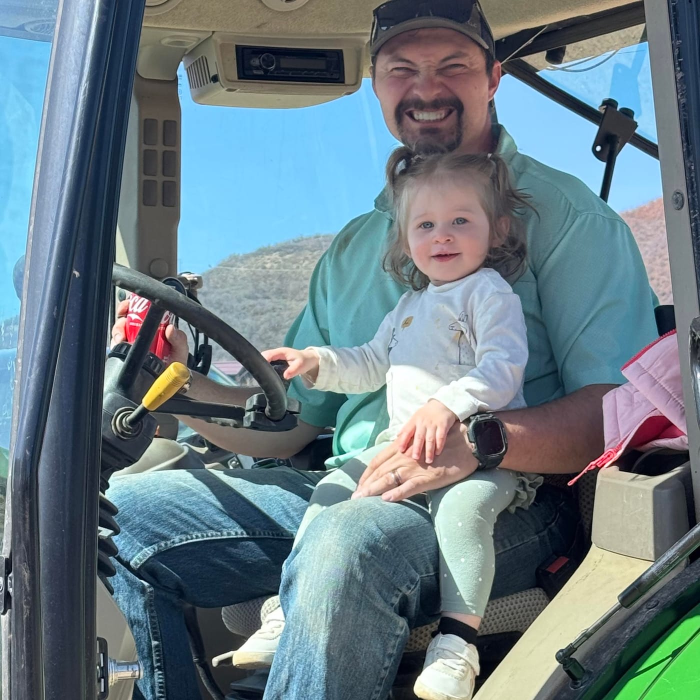 Mitch in the cab of a tractor with his daughter on his lap, both smiling.