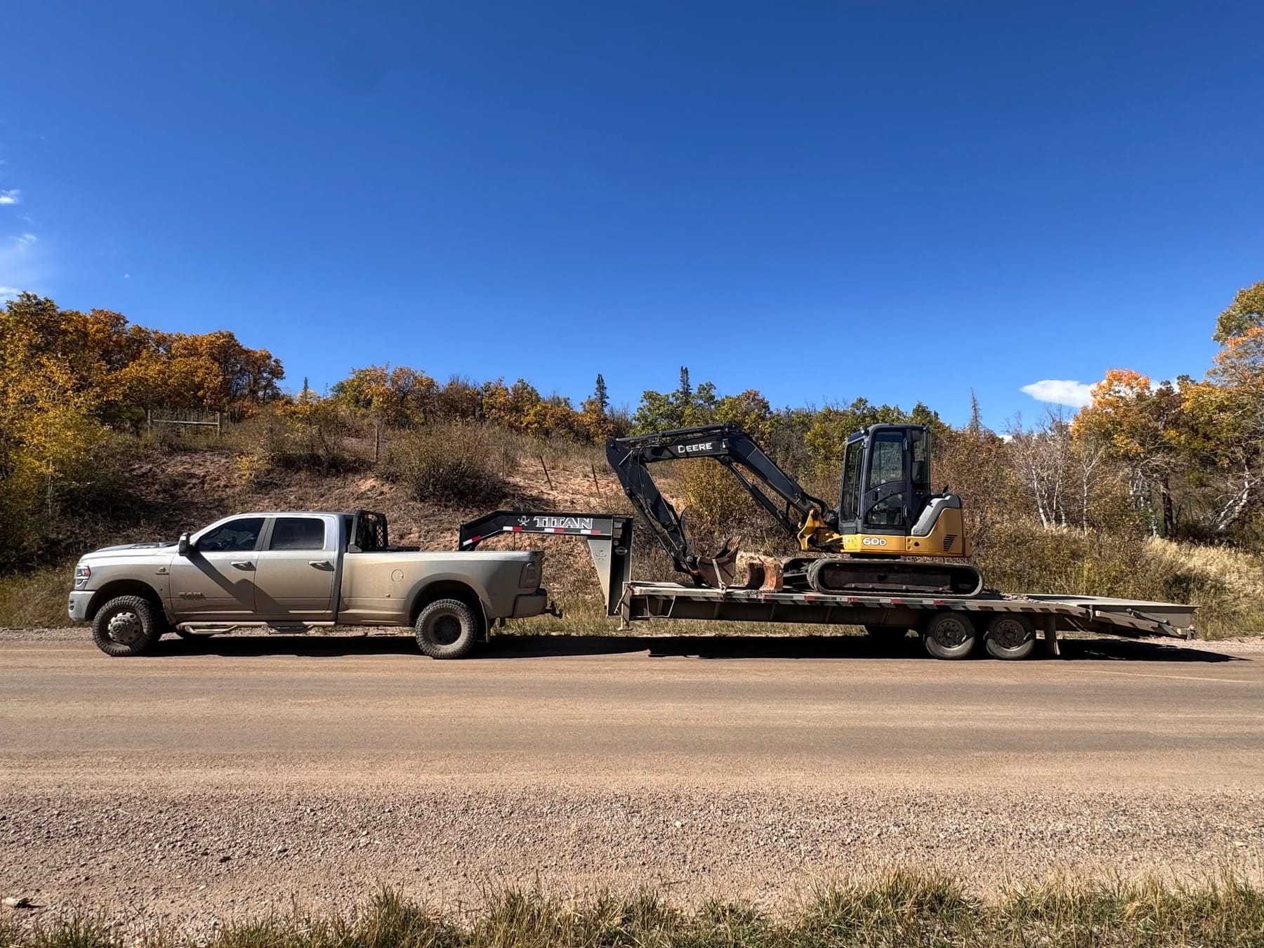 A truck and gooseneck trailer carrying a compact excavator along a gravel road lined with autumn aspens.